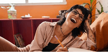 Mulher sorrindo enquanto olha para algo fora da câmera e escreve em um caderno.