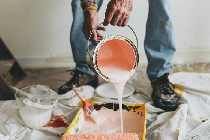 Parede amarela sendo pintada de branca. Apenas a mão do pintor, segurando o pincel, aparece na foto.