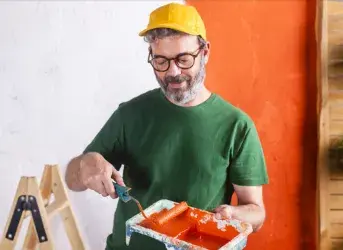 Homem segurando bandeja com tinta laranja na frente da parede que está pintando.